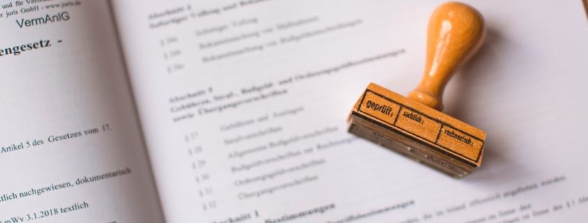 Das Bild zeigt ein offizielles Dokument mit einem Stempel, der darauf liegt. Auf dem Stempel steht das Wort "geprüft."